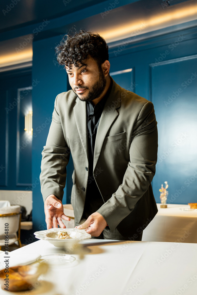 A Latino adult waiter with curly hair dressed in a stylish suit works in a modern restaurant with blue tones.The waiter is leaving a plate on the diners' table.Latin waiters concept. A Latino adult waiter with curly hair dressed in a stylish suit works in a modern restaurant with blue tones.The waiter is leaving a plate on the diners' table.Latin waiters concept.
