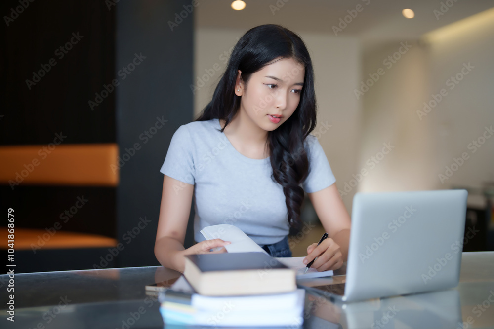 Asian teenage student woman reading a book to taking notes for assignment homework while watching lecture video online lesson on laptop to studying and learning about education knowledge in cafe Asian teenage student woman reading a book to taking notes for assignment homework while watching lecture video online lesson on laptop to studying and learning about education knowledge in cafe