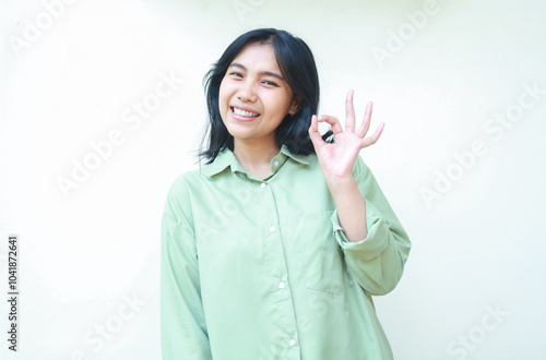 pleased satisfied asian woman with dark hair showing ok sign excelent symbols wearing green over size shirt smiling excited at camera standing over isolated white background photo