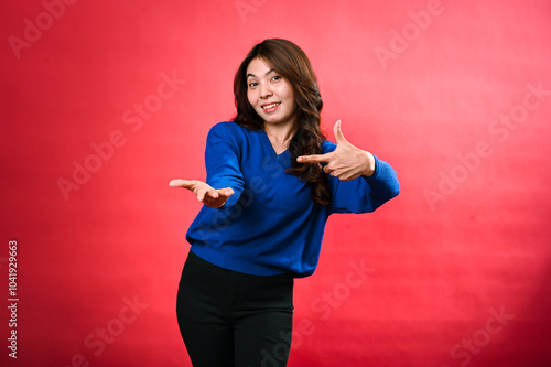 Asian woman standing against a red background, wearing a blue sweater and black pants. She is holding one hand out with her palm facing up while pointing to it with the other hand