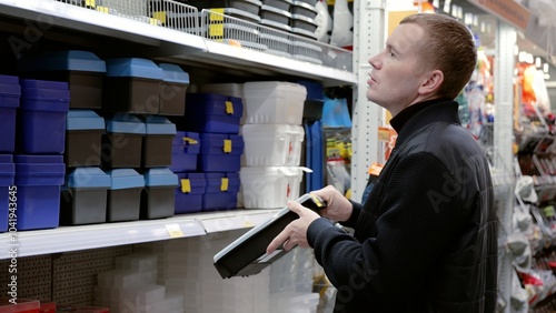 Handyman is choosing a toolbox in a hardware store, holding a black one and looking at the shelf. He is surrounded by shelves with tools and materials. A man in a hardware store choosing a tool. photo