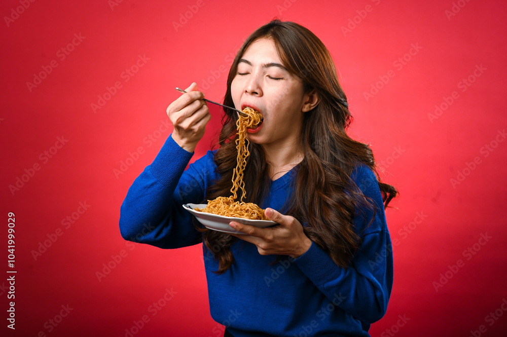 An Asian woman in a blue sweater is enjoying a plate of noodles, lifting a forkful to her mouth. She stands against a bold red background, fully focused on her delicious meal. An Asian woman in a blue sweater is enjoying a plate of noodles, lifting a forkful to her mouth. She stands against a bold red background, fully focused on her delicious meal.