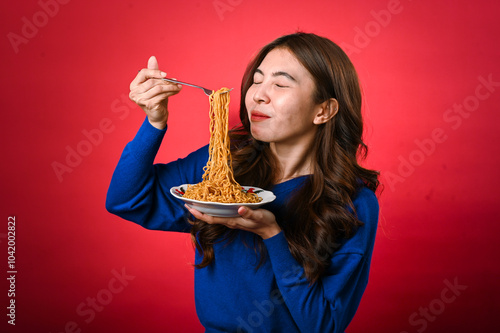 An Asian woman with long brown hair, wearing a blue sweater, lifts noodles with a fork from a plate she is holding. She smiles subtly against a red background in a playful food moment photo