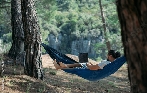 A young man is working on a laptop while lying in a hammock in the mountains.