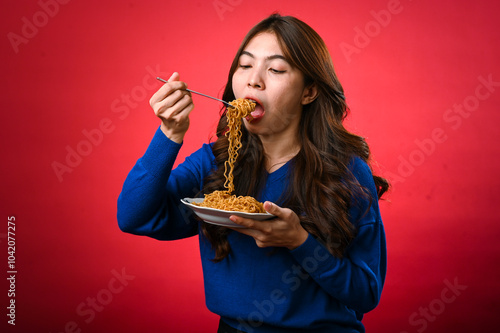 An Asian woman in a blue sweater is enjoying a plate of noodles, lifting a forkful to her mouth. She stands against a bold red background, fully focused on her delicious meal.