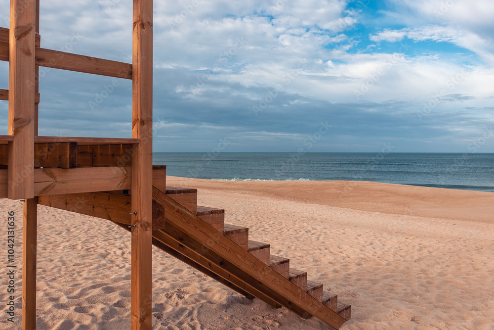 Close-up of wooden lifeguard tower stairs on a serene sandy beach in Portugal, with Atlantic Ocean in the background under a partly cloudy blue sky, Tranquil coastal landscape Close-up of wooden lifeguard tower stairs on a serene sandy beach in Portugal, with Atlantic Ocean in the background under a partly cloudy blue sky, Tranquil coastal landscape