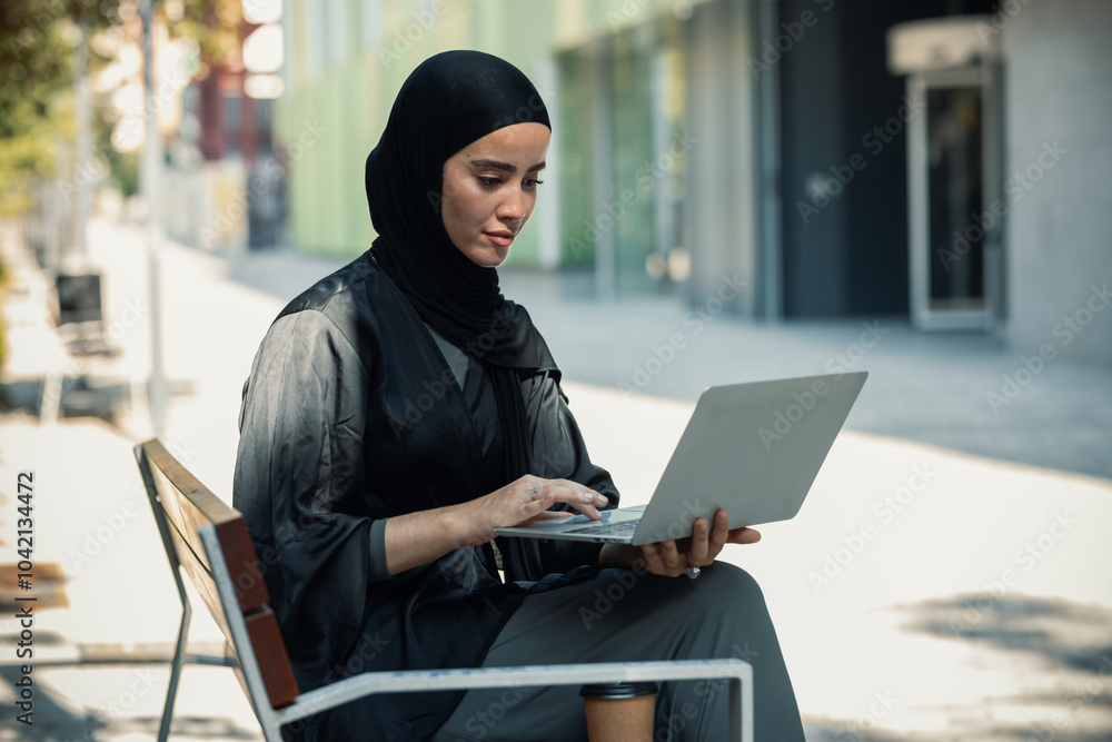 Pretty woman with hijab working with laptop sitting on a bench in the city Pretty woman with hijab working with laptop sitting on a bench in the city