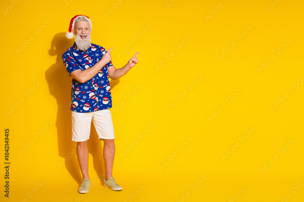 Full size photo of funny stylish santa indicate fingers empty space christmas print shirt isolated on yellow color background Full size photo of funny stylish santa indicate fingers empty space christmas print shirt isolated on yellow color background