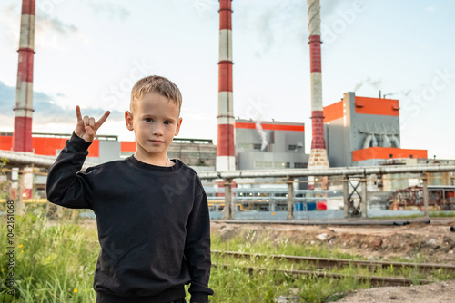 A boy stands against the backdrop of a factory.