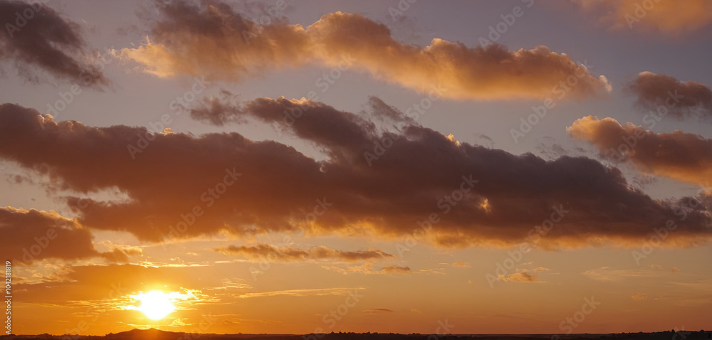 Naklejka premium The sun sets behind rolling hills, casting a warm glow across the horizon with clouds softly illuminated in shades of orange and pink during the evening