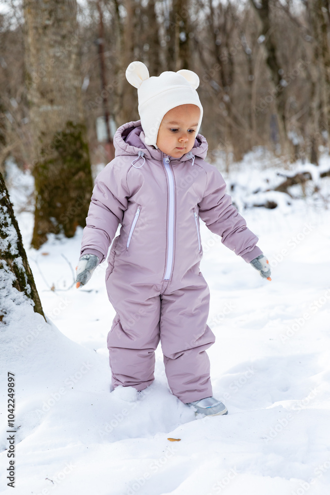Side view toddler girl in light pink jumpsuit and a white fluffy hat walking in winter park. Children games in the snowy forest. Family winter vacation with a child. Vertical photo Side view toddler girl in light pink jumpsuit and a white fluffy hat walking in winter park. Children games in the snowy forest. Family winter vacation with a child. Vertical photo