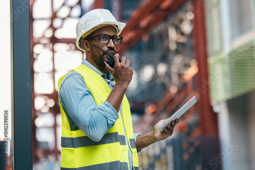 Attractive man with worker's helmet talking on the walkie talkie while passing list with digital tablet near a construction