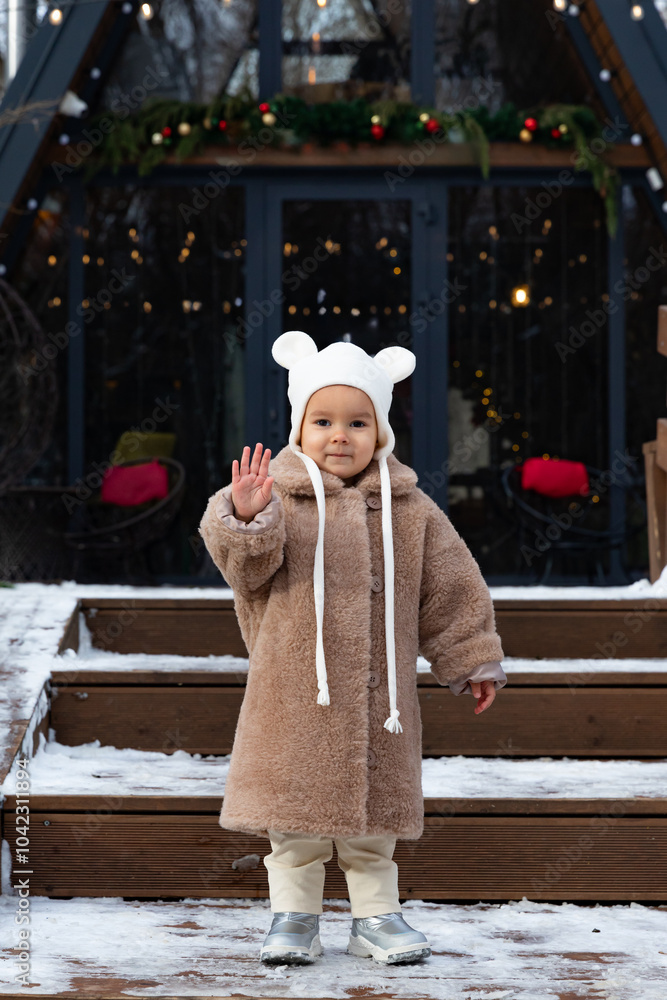 Obraz premium Little girl in a brown fashionable fur coat on the background of festive decorated a-frame house. Family vacation time. High quality vertical photo. Winter outdoor activity