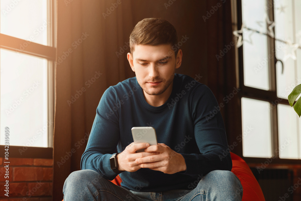 Millennial Guy Using Smartphone Texting Sitting In Beanbag Chair In Modern Loft Interior Indoor. Selective Focus Millennial Guy Using Smartphone Texting Sitting In Beanbag Chair In Modern Loft Interior Indoor. Selective Focus