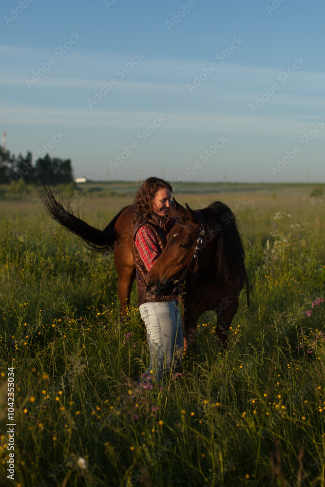 Young beautiful woman in brown vest and jeans holding reins and hugging brown horse in field at dawn