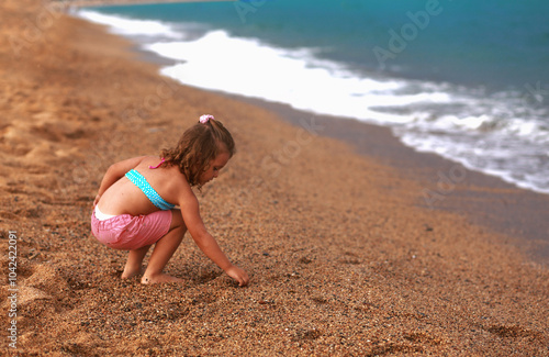 Beautiful fun happy kid girl sitting in summer clothing on the yellow sand and playong with stones of the beach near blue sea and looking on water background. Happy photo