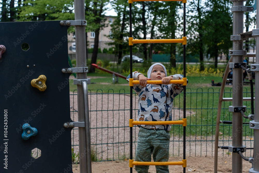 Little boy in a dinosaur hoodie climbing a yellow rope ladder on a playground on an overcast day. Concept of childhood development, outdoor activities, and building physical strength. . High quality Little boy in a dinosaur hoodie climbing a yellow rope ladder on a playground on an overcast day. Concept of childhood development, outdoor activities, and building physical strength. . High quality