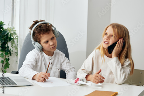 Happy Caucasian boy and girl studying at home with their laptops, headphones, and tablets They are engaged in online learning, sitting at a table in the living room Their concentration and connection