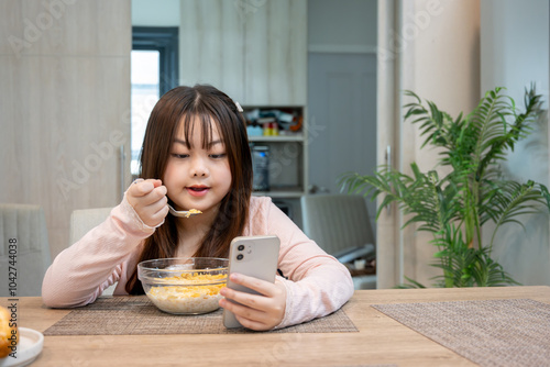 Asian girl enjoys watching cartoons on her smartphone while having breakfast and eating cereal at the dining table.