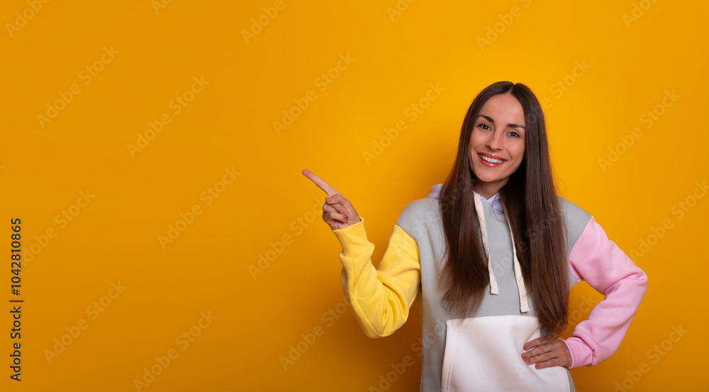 A smiling young woman stands against a vibrant yellow background, wearing hoodie. She points to her left with one hand, directing attention, reflecting a cheerful and engaging vibe. A smiling young woman stands against a vibrant yellow background, wearing hoodie. She points to her left with one hand, directing attention, reflecting a cheerful and engaging vibe.