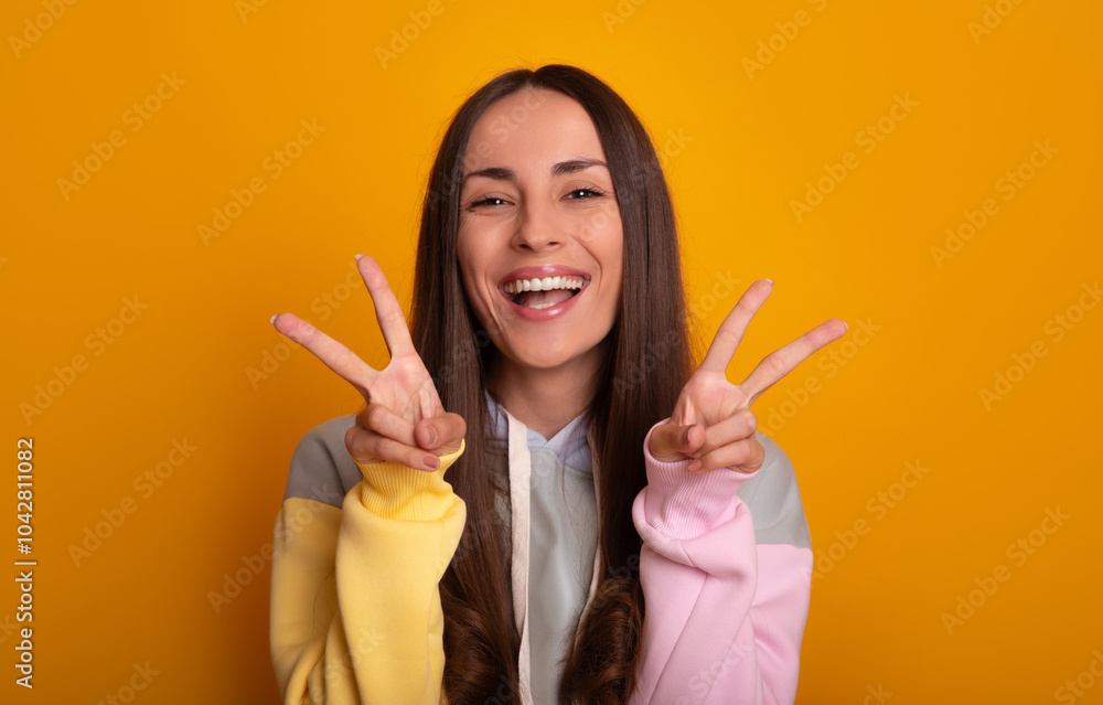 Cheerful young woman posing against vibrant yellow background, flashing double peace signs with both hands. She wears casual, colorful hoodie and smiles broadly, radiating positivity and fun energy Cheerful young woman posing against vibrant yellow background, flashing double peace signs with both hands. She wears casual, colorful hoodie and smiles broadly, radiating positivity and fun energy