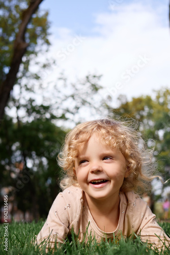 Happy Caucasian little girl with wavy blond hair smiling with joy while playing outdoors in park looking adorable and cute enjoying beautiful green park nature