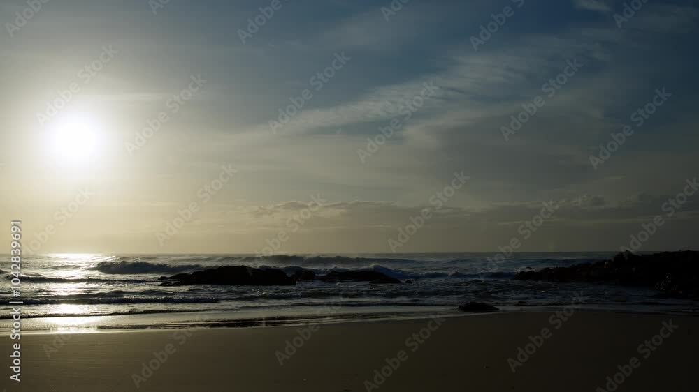 A wide shot captures waves rolling past rocks, as the sun in the background casts a soft glare across the water and glistening wet sand. A wide shot captures waves rolling past rocks, as the sun in the background casts a soft glare across the water and glistening wet sand.