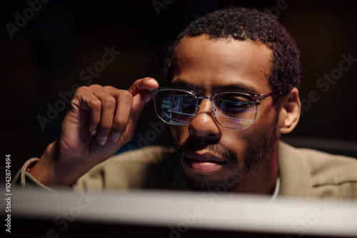 Fototapeta Man sitting at a workstation adjusting his glasses while focusing intently on a