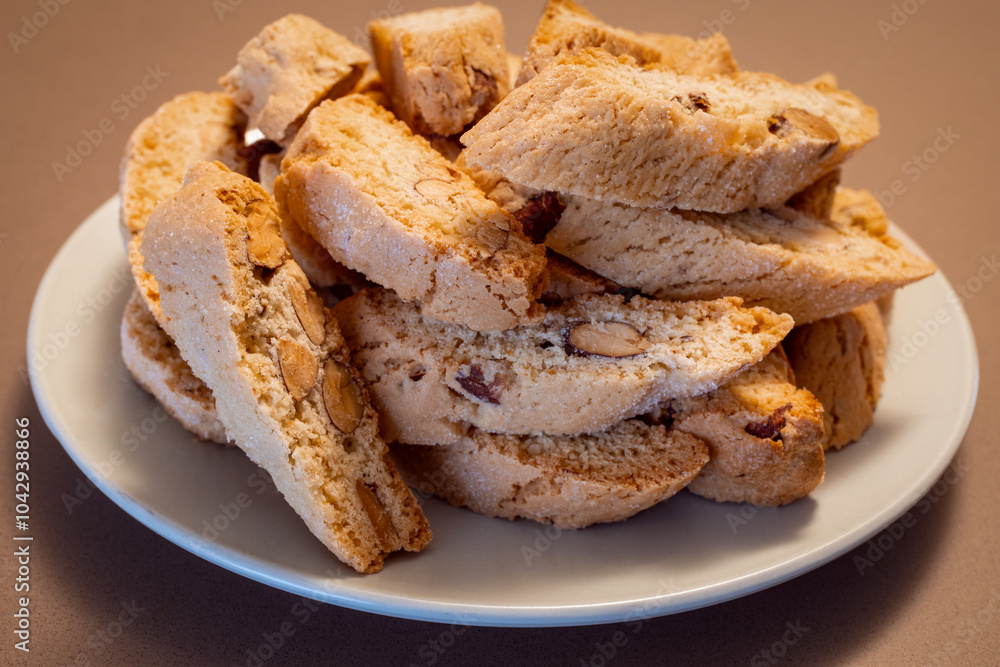 A plate of Italian almond cantucci cookies piled on a brown surface The golden-brown biscotti with visible almond pieces highlight their crunchy texture and nutty flavor A plate of Italian almond cantucci cookies piled on a brown surface The golden-brown biscotti with visible almond pieces highlight their crunchy texture and nutty flavor