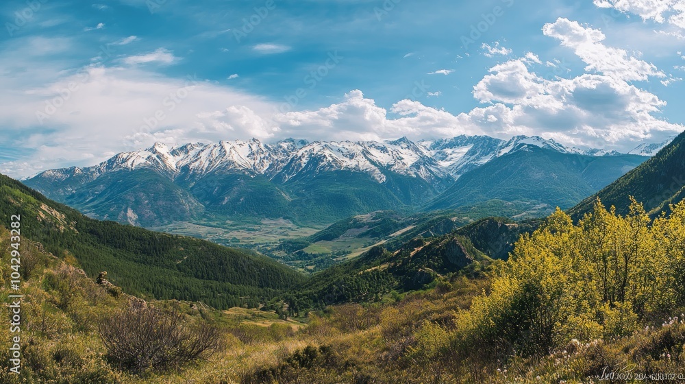 Fototapeta premium A panoramic view of a snow-capped mountain range, with green valleys and a blue sky with white clouds.
