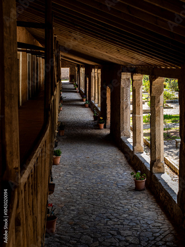 Long, shaded Porxets corridor at Lluc Monastery in Mallorca, with rustic stone columns on one side and potted plants lining the cobbled path, reflecting Mediterranean culture and historic architecture photo