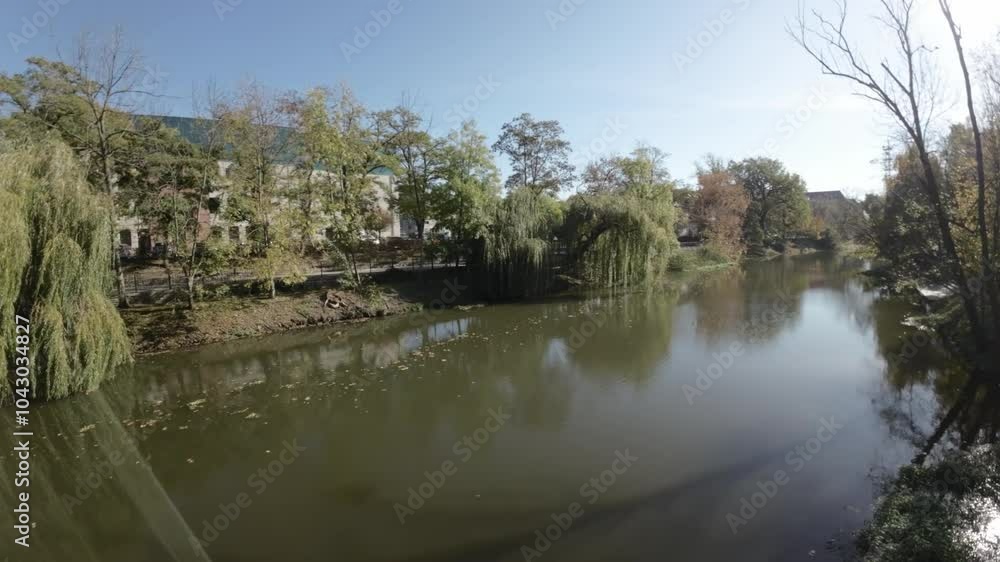 The video begins with a first-person view FPV of a drone flying over a river in autumn. The drone flies along the river, passing under bridges and through trees. The water is a deep green color The video begins with a first-person view FPV of a drone flying over a river in autumn. The drone flies along the river, passing under bridges and through trees. The water is a deep green color