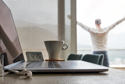 Laptop with headphones sits on a table with a blurred background of a person standing by a window. Home office concept