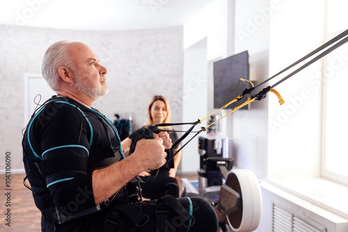 Elderly male athletic in electrical muscle stimulation suit uses trx system for his workout with personal trainer photo