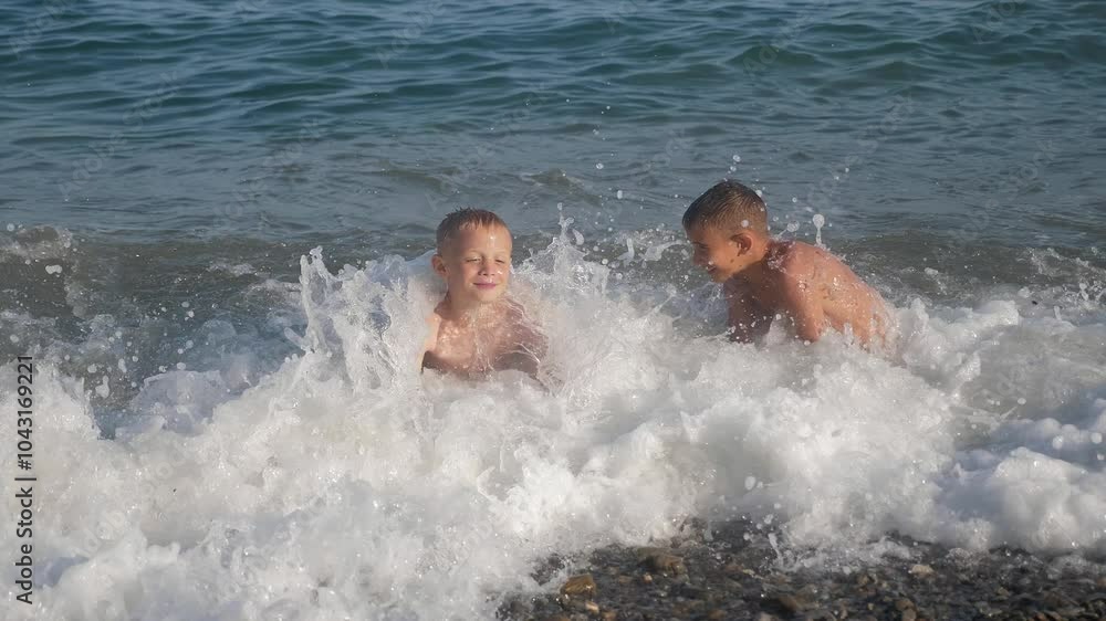 Two happy teenage boys are playing on the beach with big waves in stormy weather. Strong waves cover two boys who are sitting on the shore. Two happy teenage boys are playing on the beach with big waves in stormy weather. Strong waves cover two boys who are sitting on the shore.