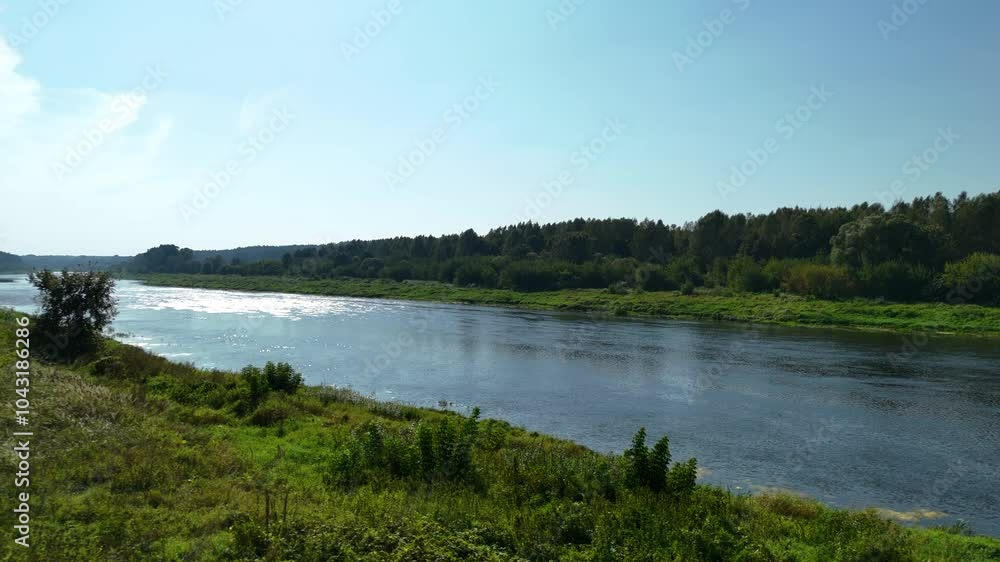 4K. A calm Daugava river reflecting the sunlight as it flows through a peaceful, green Latgale landscape. Lush vegetation lines the riverbanks under a clear blue sky, creating a serene natural scene. 4K. A calm Daugava river reflecting the sunlight as it flows through a peaceful, green Latgale landscape. Lush vegetation lines the riverbanks under a clear blue sky, creating a serene natural scene.
