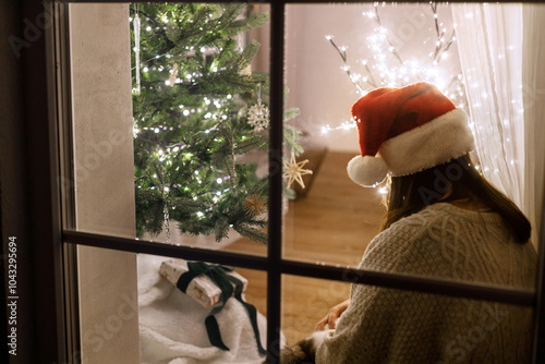 Beautiful woman in santa hat decorating stylish christmas tree in window on background of festive living room with lights. Atmospheric christmas eve at home. Merry Christmas! Holiday magical scene