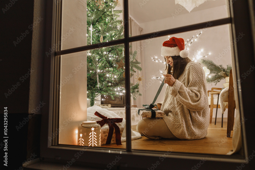 Obraz premium Merry Christmas! Beautiful woman in santa hat opening stylish christmas gift under christmas tree in window on background of festive living room with lights. Atmospheric christmas eve at home