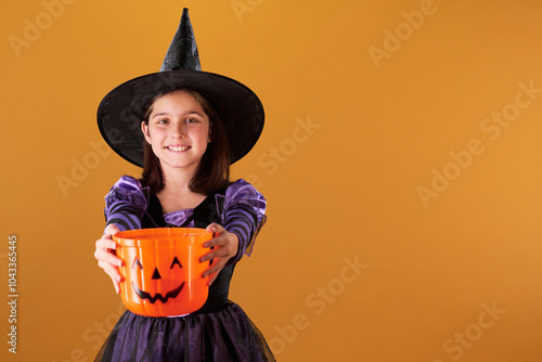 Smiling little girl in fancy witch costume holding out Halloween pumpkin bucket for trick or treating in isolation photo