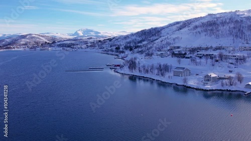 Snow covered mountain range on coastline in winter, Norway. Surroundings of town Tromso. Panoramic aerial view landscape of nordic snow cowered mountains, houses and ocean. Troms county, Fjordgard