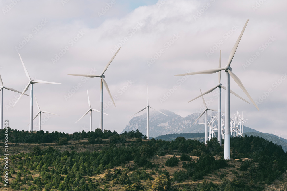 A cluster of towering wind turbines stands firm at the base of a mountain range on a partially cloudy day, representing clean energy and modern sustainability solutions in Navarra Spain A cluster of towering wind turbines stands firm at the base of a mountain range on a partially cloudy day, representing clean energy and modern sustainability solutions in Navarra Spain