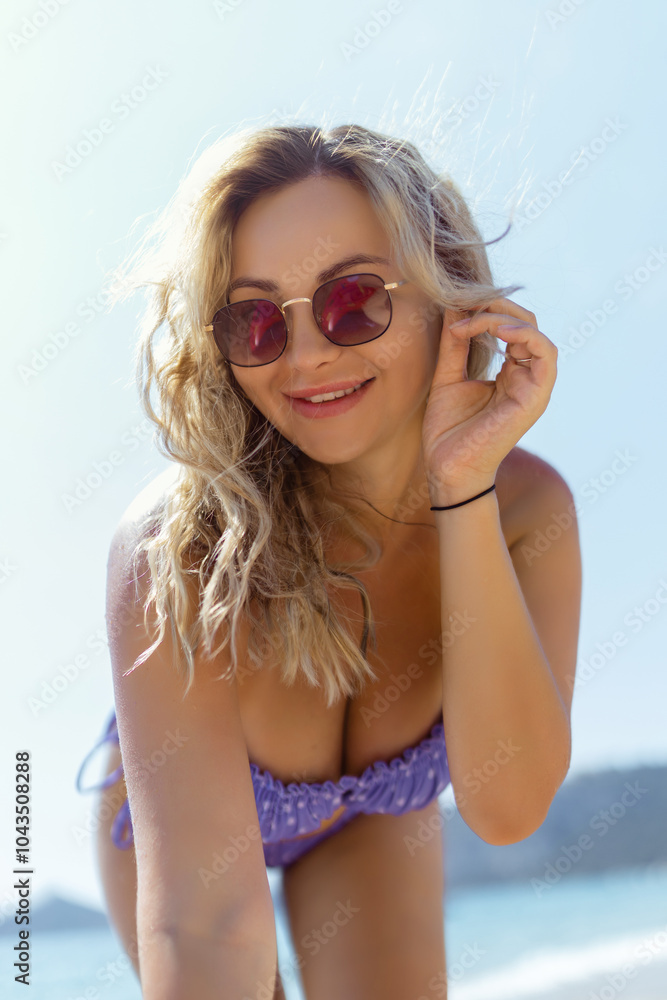 A beautiful girl in sunglasses on the beach looks at the camera, leaning towards it. Sexy blonde in a swimsuit on the seashore A beautiful girl in sunglasses on the beach looks at the camera, leaning towards it. Sexy blonde in a swimsuit on the seashore