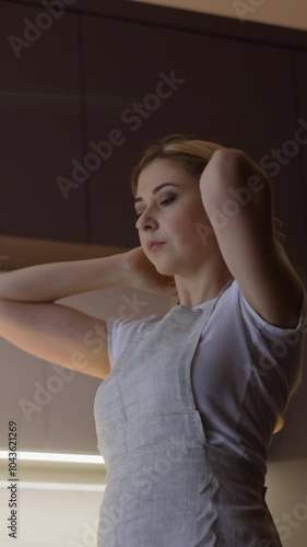 Pretty woman in light grey apron adjusts long fair hair ready to cook delicious healthy dinner in contemporary spacious kitchen slow motion photo