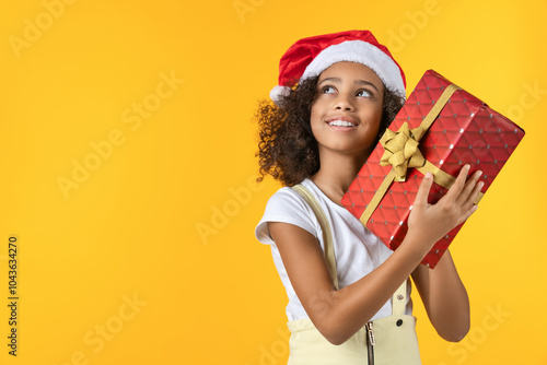 Happy african girl in Christmas hat holding big red present gift box isolated on yellow background looking aside up. Gift sharing for New Year holiday concept copy free space