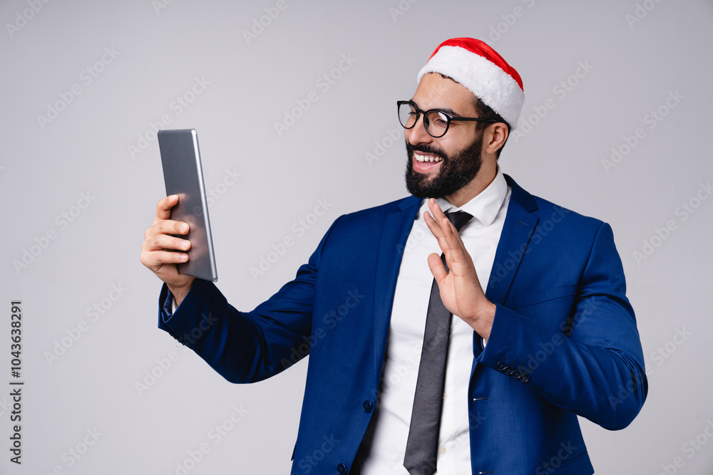 Videocall conference meeting. Arabian businessman in Santa Claus red hat using digital tablet for online shopping for Christmas New Year, texting, surfing social media on grey background Videocall conference meeting. Arabian businessman in Santa Claus red hat using digital tablet for online shopping for Christmas New Year, texting, surfing social media on grey background