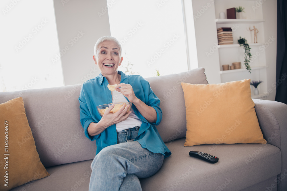 Photo of nice aged lady watch tv eat chips wear blue shirt bright interior apartment indoors Photo of nice aged lady watch tv eat chips wear blue shirt bright interior apartment indoors