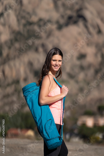 Smiling woman in pink tank top carrying a blue yoga mat bag, walking confidently outdoors with mountains.