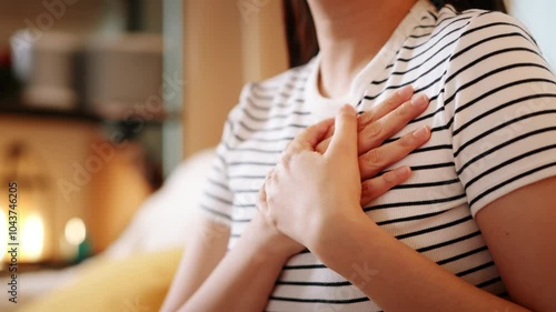 A young woman in a striped shirt sits comfortably on a sofa with hands over her heart, representing self-compassion and mindfulness in a peaceful indoor environment.