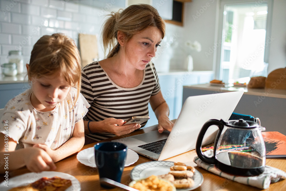 Mother working on laptop while family has breakfast in kitchen Mother working on laptop while family has breakfast in kitchen