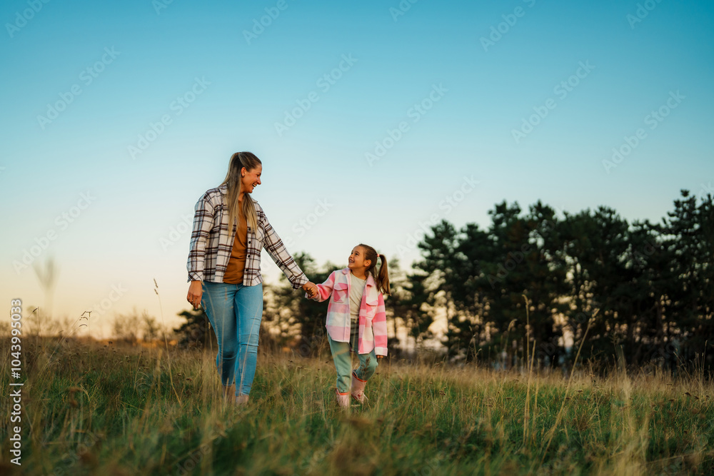 A mother and daughter walking hand in hand through a grassy field at sunset, with trees in the background. A mother and daughter walking hand in hand through a grassy field at sunset, with trees in the background.